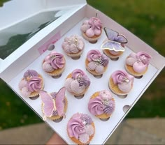 a person holding up a box of cupcakes with pink frosting and butterflies on them