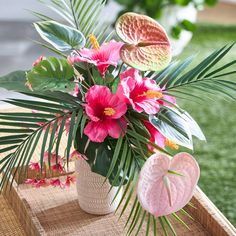 pink flowers and palm leaves in a vase on a wooden table with grass behind it