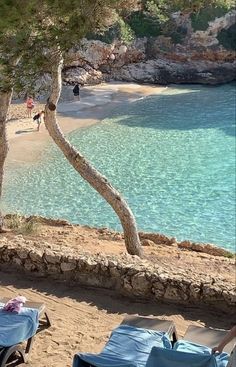 two lounge chairs sitting on the beach next to some trees and people walking in the water