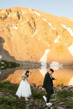 a bride and groom are walking by the water in front of a mountain with snow on it