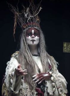 a woman with white makeup and long hair wearing a headdress in front of a black background