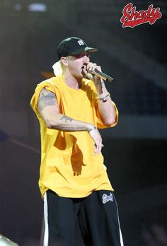 a male in a yellow shirt and black pants holding a microphone to his mouth while standing on stage