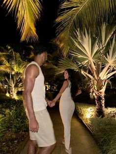 a man and woman holding hands while walking down a path in the night with palm trees behind them