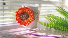 an orange crocheted flower sits on a coffee cup next to a green plant