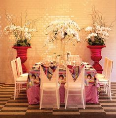 the table is set up with white chairs and pink cloths on it, along with flowers in large red vases