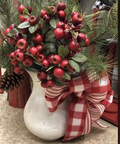 a white vase filled with red berries and greenery on top of a counter next to a pine cone