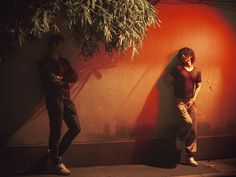 two men standing next to each other in front of a red wall and green tree