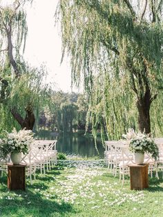 an outdoor ceremony set up with white chairs and flowers on the grass, under willow trees