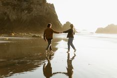 two people are walking on the beach holding hands and looking out at the ocean with cliffs in the background