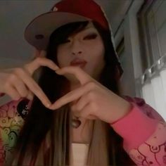 a girl making a heart sign with her hands while wearing a baseball cap and pink shirt