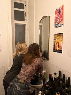 two women sitting on a toilet in front of a mirror filled with bottles and wine