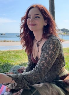 a woman with red hair sitting on the grass by the beach and smiling at the camera