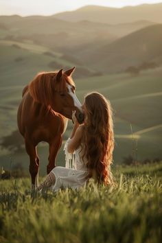 a woman sitting in the grass next to a brown horse and kissing it's face