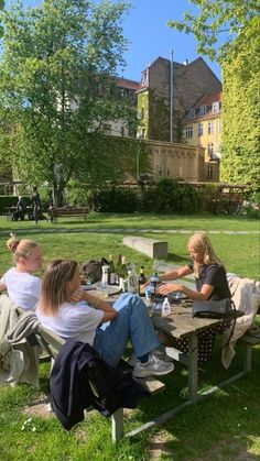 three people sitting at a picnic table in the park eating and drinking wine while another person looks on