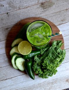 a wooden plate topped with cucumbers, limes and broccoli next to a glass of green juice