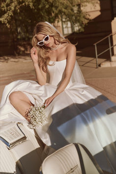 a woman sitting on the back of a car wearing sunglasses and a wedding dress with flowers in her hand