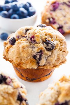 muffins with blueberries and crumbs are on a white surface next to a bowl of berries