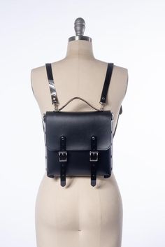 The Blackout Harness Mini Backpack is a medium-sized bag for running around and doing all the things. The black vegan leather and shiny black PVC pair nicely together for a pleasing black-on-black texture contrast. Features two buckle front closures, detachable shoulder straps can convert to a shoulder strap by attaching to the side d-rings, and a handle on top.Silver toned hardware.BAG DIMENSIONSHeight:  9.75 inches (20.32 cm)Width:  8.75 inches (21.59 cm)Depth: 2 inches (10.16 cm)All pieces ar Black Satchel With Hardware Details, Gothic Style Standard Backpack For Travel, Gothic Standard Backpack For Travel, Punk Style School Satchel Bag, Gothic Black Backpack Bag, Black Leather Backpack With Gunmetal Hardware For Daily Use, Punk Style Standard Backpack For Everyday Use, Gothic Standard Backpack For Everyday Use, Edgy Standard Backpack For Everyday Use