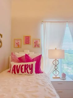 a white bed topped with pillows next to a window covered in pink and white curtains