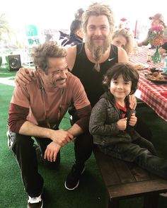 a man sitting next to a little boy on top of a wooden bench in front of a picnic table