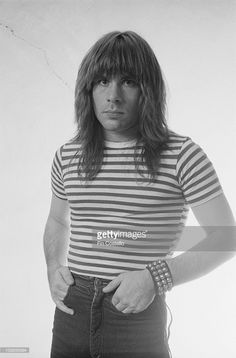 a man with long hair standing in front of a white wall wearing a striped shirt