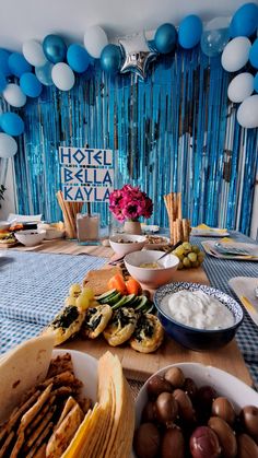 a table topped with plates and bowls filled with food next to blue balloons hanging from the ceiling