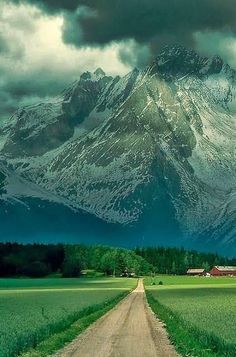 a dirt road in front of a snow covered mountain with green grass on both sides