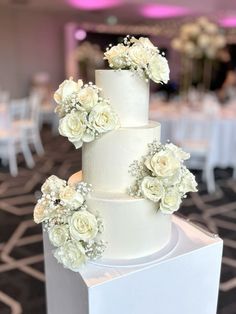 a three tiered white wedding cake with flowers on the top and sides, in a banquet room