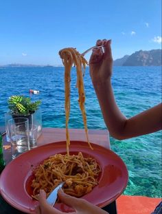 a person is eating spaghetti on a red plate near the ocean and blue water with mountains in the background