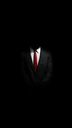 a man in a suit and red tie standing in the dark with his arms crossed