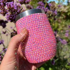 a hand holding a pink cup in front of some purple and white flowers with glitter on it