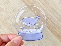 a hand holding a snow globe with a cartoon whale inside it on top of a wooden table