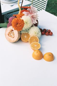 an arrangement of flowers, oranges and other fruit on a white tablecloth with chairs in the background