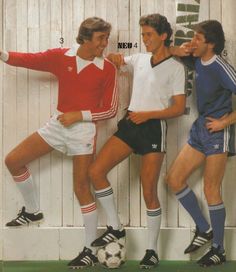 three men standing next to each other near a wall with soccer balls on it and one man leaning against the wall