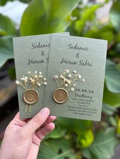 two wedding cards with flowers on them are held in front of some plants and leaves