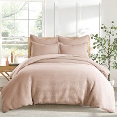 a bed with pink comforter and pillows in a white room next to a potted plant