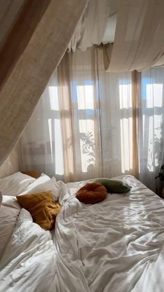 an unmade bed with white sheets and pillows in front of two windows that have sheer drapes on them