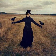 a woman standing in a field with two birds on her arm and wearing a top hat