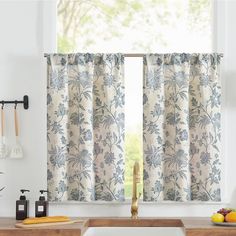 a kitchen window with blue and white curtains in front of an open sink, cutting board, utensils and fruit on the counter