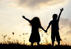 two children are holding hands and standing in the grass at sunset with their arms outstretched