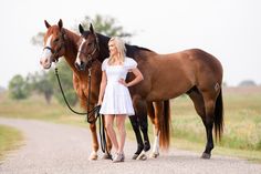 a woman standing next to two brown horses on a country road with grass and trees in the background