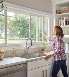 a woman standing in front of a kitchen sink next to a window with lots of windows