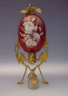 an ornate red and gold vase with white flowers on it's stand, against a gray background