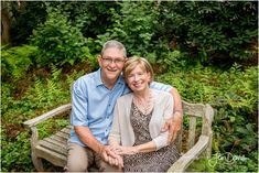 an older man and woman sitting on a bench in front of some bushes, smiling at the camera