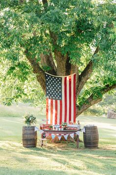 an american flag draped over a picnic table under a large tree with two wooden barrels in the foreground