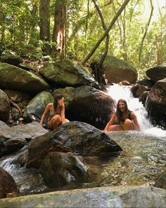 two women in bathing suits sitting on rocks next to a waterfall with water running over them