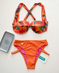 Two-piece bikini set laid flat, featuring an abstract, fiery print on the top and a solid orange bottom, with a blue and white patterned smartphone case and brand tag to the side. Tropical Style Tankini With Adjustable Straps For Beach Season, Tropical Tankini With Adjustable Straps For Vacation, Vibrant Orange Swimwear For The Beach, Vibrant Orange Swimwear For Beach, Tropical Swimwear With Adjustable Straps For Beach Party, Tropical Beach Swimwear With Adjustable Straps, Vibrant Tropical Print Swimwear For The Beach, Vibrant Swimwear With Tropical Print For Beach, Vibrant Tropical Print Swimwear For Beach