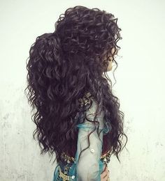 a woman with long dark curly hair standing in front of a white wall wearing a blue dress