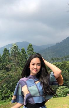 a woman posing for a photo in front of some trees and mountains with her hands on her head