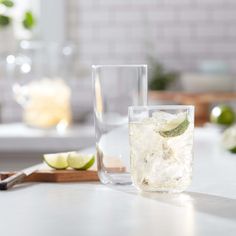 two glasses filled with ice and limes sitting on a table next to a cutting board